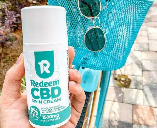 Hand holding a CBD Pain Cream bottle against the backdrop of a teal-colored bicycle with a pair of sunglasses resting in the basket.