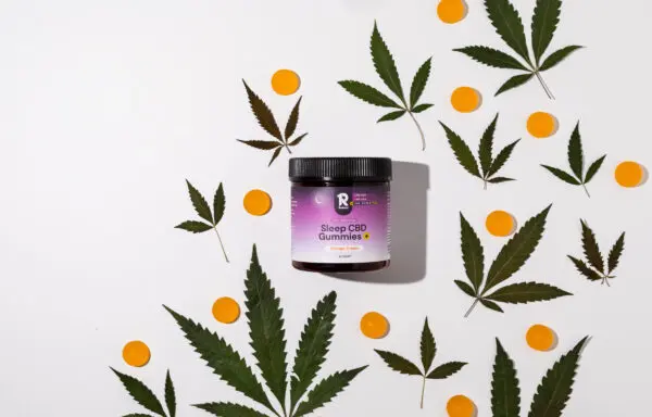 A jar of Sleep CBD Gummies+ (Orange Cream flavor) surrounded by cannabis leaves and orange gummies