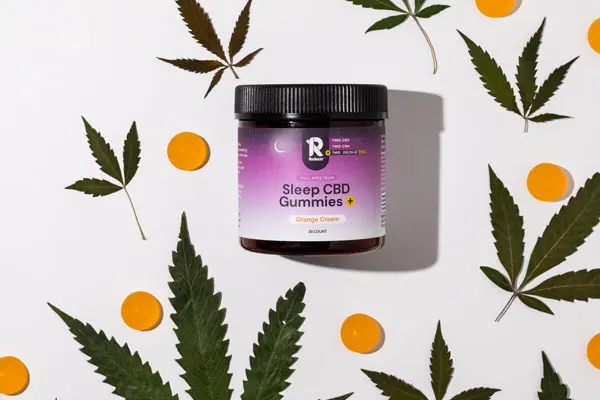 A jar of Sleep CBD Gummies in Orange Cream flavor, surrounded by cannabis leaves and orange gummies.